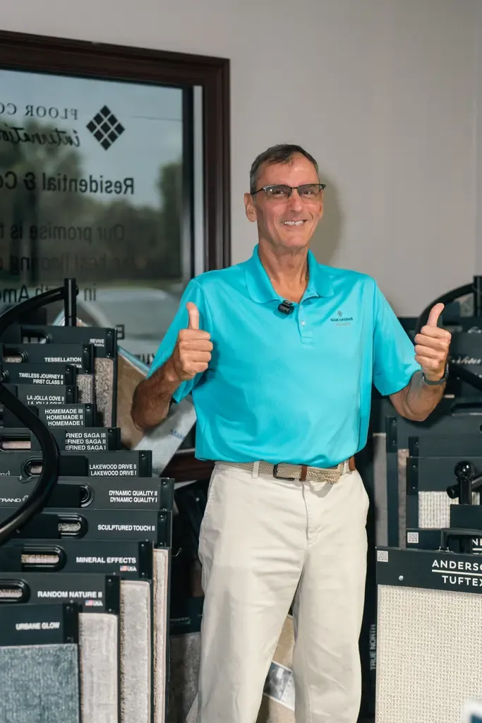 Man wearing glasses and turquoise polo gives a thumbs up in a carpet showroom. Displays of carpet samples surround him, creating a welcoming vibe.