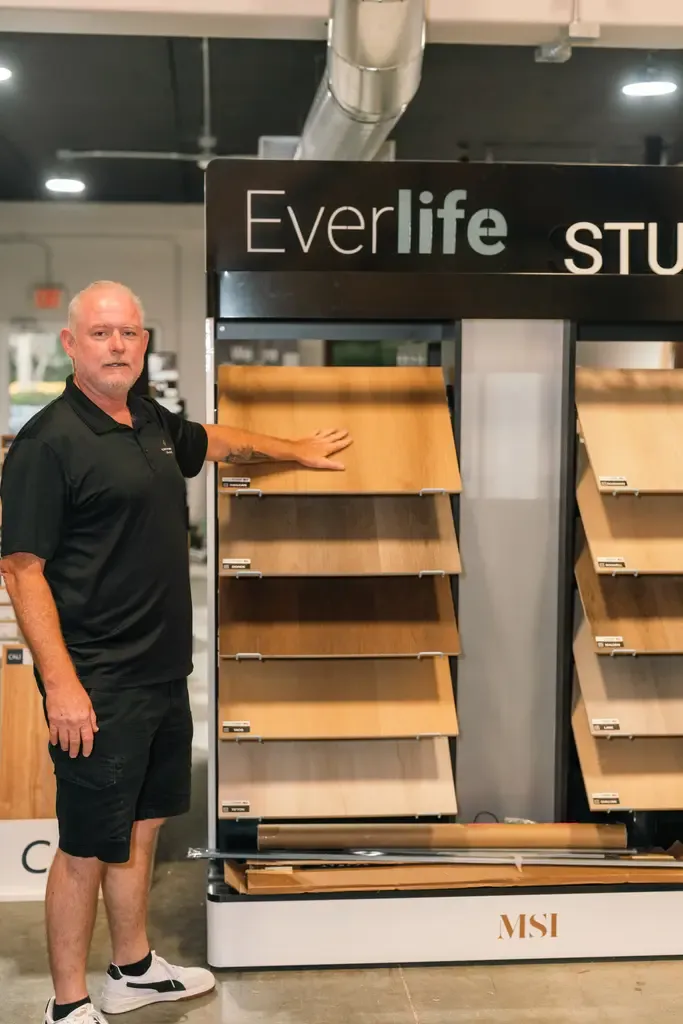 A man in a black polo and shorts stands in a showroom next to a display of wooden flooring samples. The display sign reads "Everlife Studio."