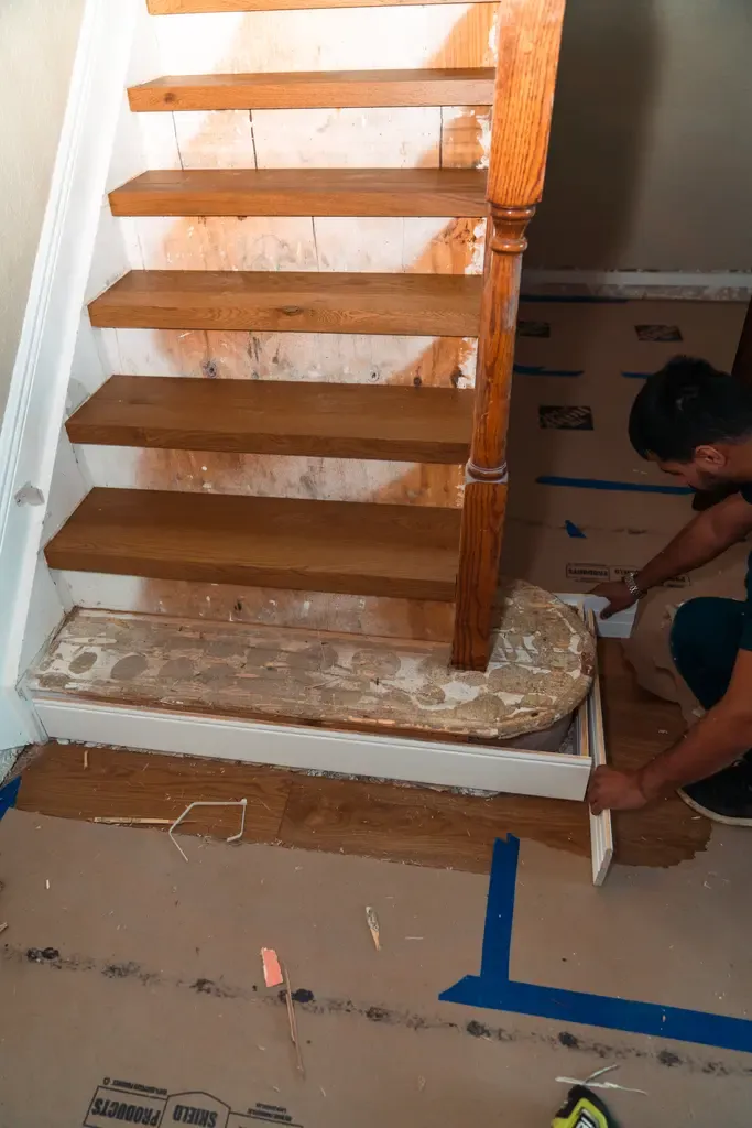 A person in Wellington installs a new wooden stair tread and riser at the bottom of a staircase; tools, wood flooring materials, and protective floor covering are visible.