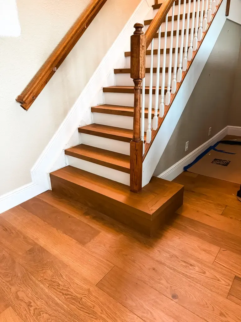 Wooden staircase with white risers and spindles, brown handrail, and elegant wood flooring; a section of the floor near the stairs is taped for protection, reflecting classic Wellington, FL style.