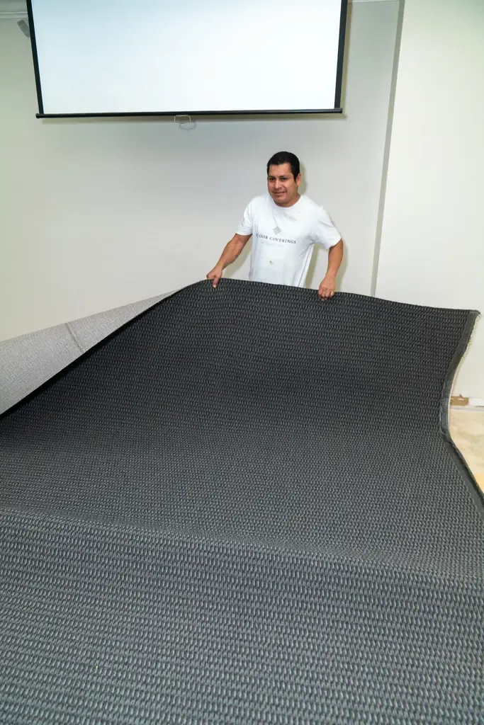 Man in white shirt smiles while unrolling a large black textured carpet in a room with white walls and a ceiling-mounted projector screen.