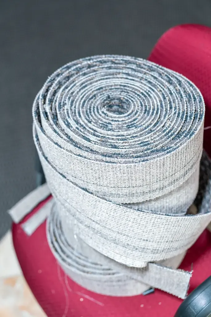 A large, tightly rolled coil of grey jute webbing sits on a red cushioned chair, set against a dark, textured background. The scene is industrial.