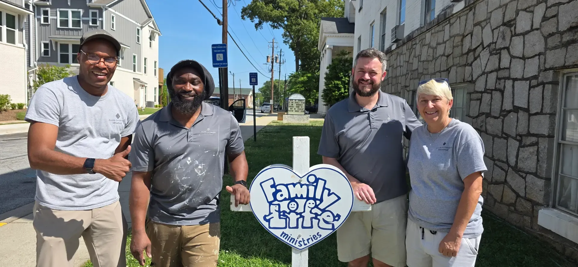A group of four people wearing grey shirts poses next to a sign, showcasing a sense of camaraderie and teamwork.