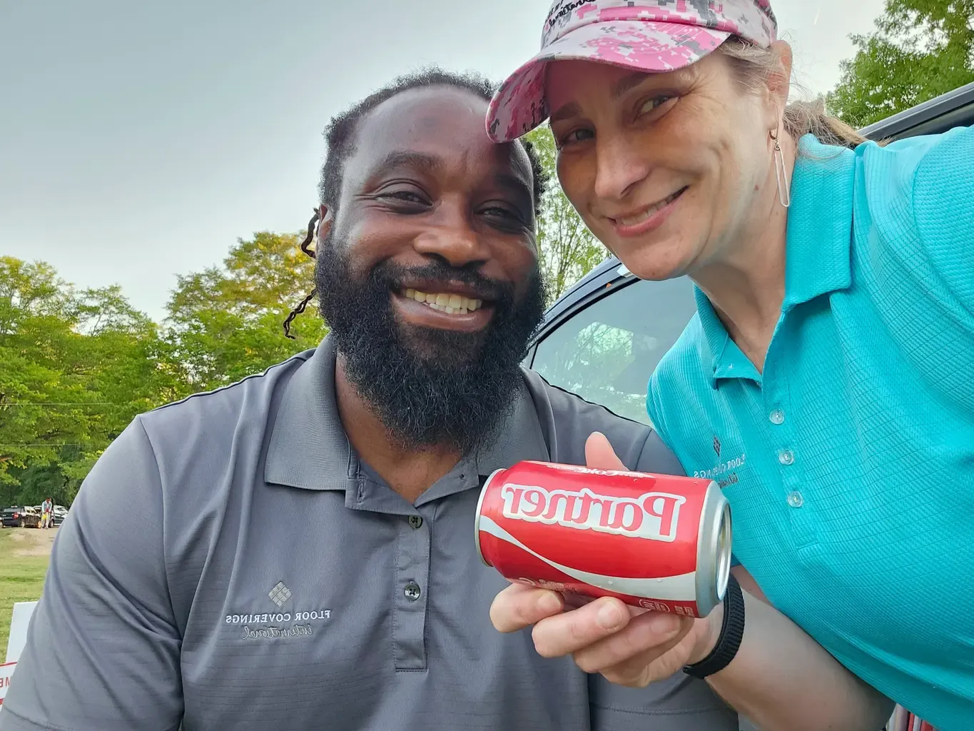 A couple stands side by side, each holding a Coke can, smiling for the camera in a cheerful setting.