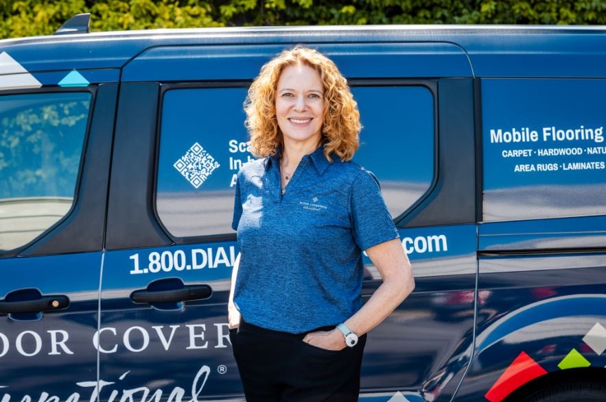A woman with curly hair wearing a blue polo shirt stands in front of a Floor Coverings International van